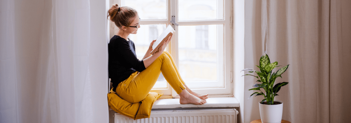 En kvinna i svart tröja och gula byxor sitter i ett fönster och läser i ljuset från solen