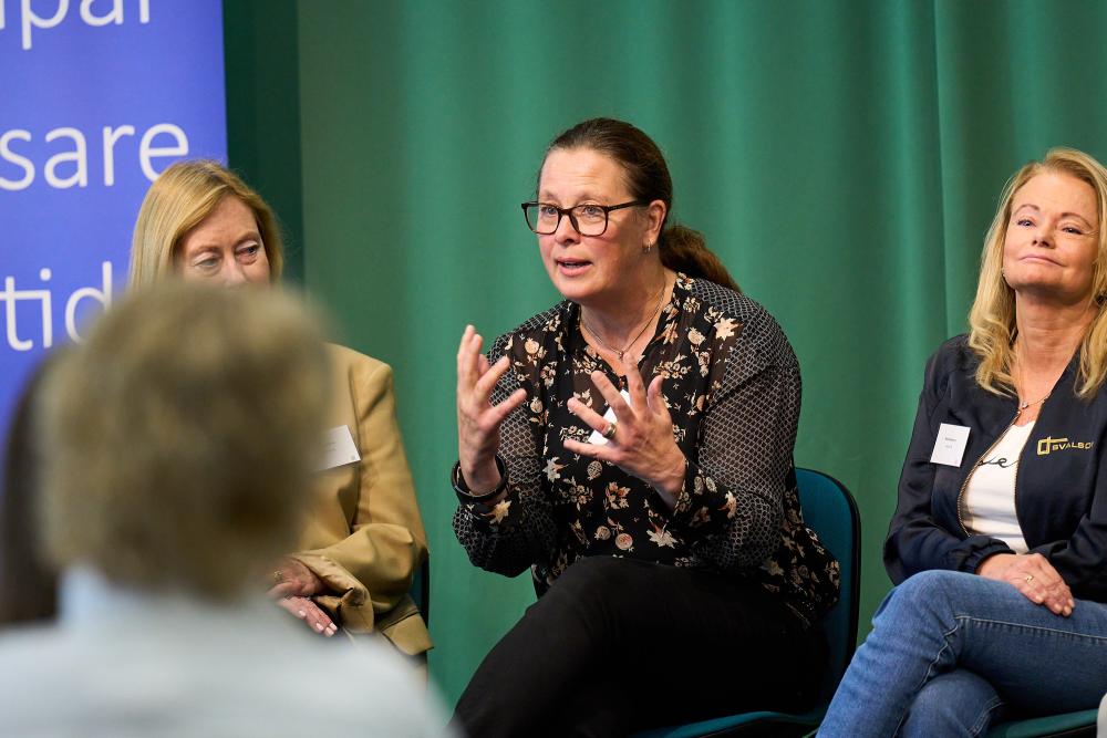 Kvinna i glasögon och blommig tröja gestikulerar med händerna på en scen med grönt tyg i bakgrunden