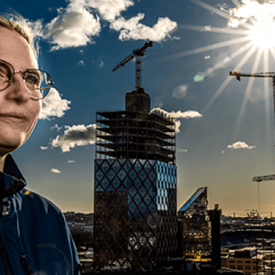 En kvinna till vänster i bild i förgrunden. I bakgrunden lyser solen mot blå himmel över en byggnad med glasfasad under konstruktion Foto: Sören Håkanlind.