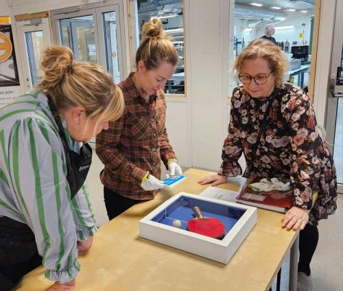 Tre kvinnor står lutade över bord och granskar inramning