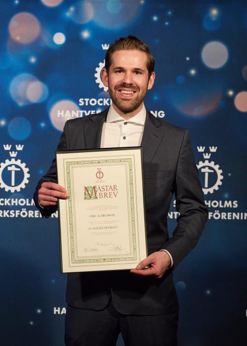 Man i kostym håller i inramning av mästarbrev i glasmästeriyrket