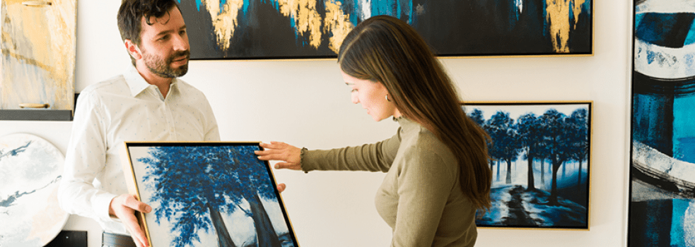 En kvinna betraktar en målning i ram som visas upp av en man. Bakom dem en vägg med tavlor.