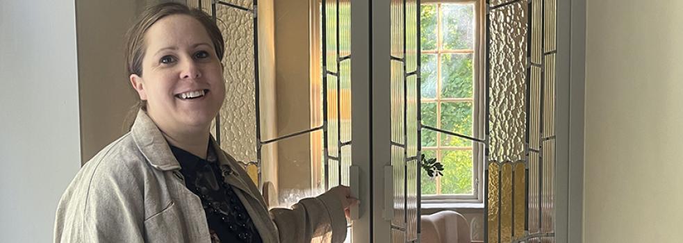 Leende kvinna i beige jacka framför pardörrar med blyglasinfattningar