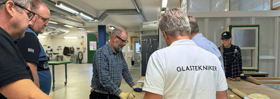 Yrkeslärare står runtom arbetsbord i verkstad och tittar på ett arbetsmoment med glas