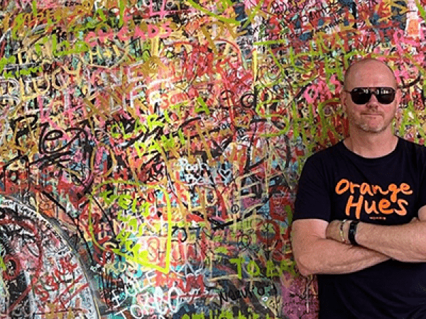 Man i svart -tshirt och solglasögon står framför en vägg med graffiti 