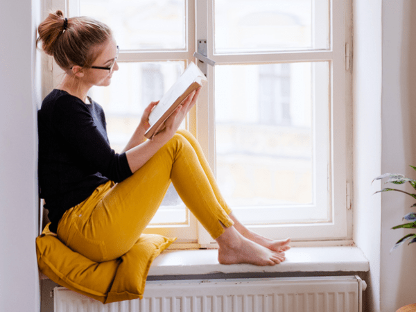 En kvinna i svart tröja och gula byxor sitter i ett fönster och läser i ljuset från solen