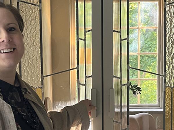 Leende kvinna i beige jacka framför pardörrar med blyglasinfattningar