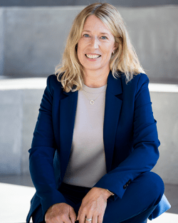 Blond kvinna i blå kostym sitter ned och tittar leendes in i kameran