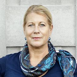 Blond kvinna i mönstrad sjal och blå topp tittar in i kameran